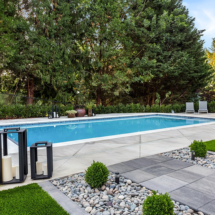 A serene backyard features a rectangular swimming pool, surrounded by lush trees, pebbles, and modern patio furniture, creating a tranquil outdoor setting.
