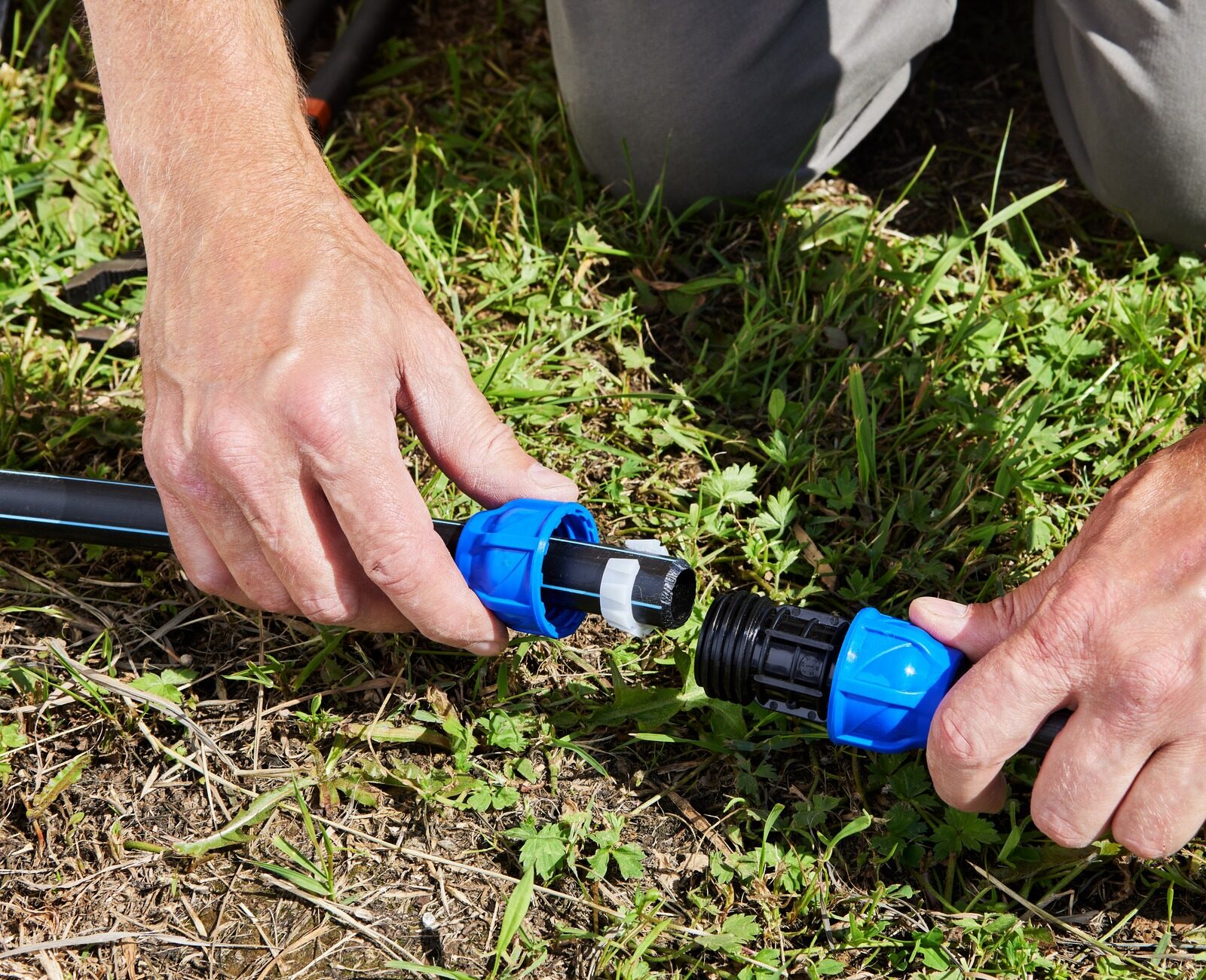 A person assembling a black hose with blue connectors on grassy ground, using both hands for a secure connection.