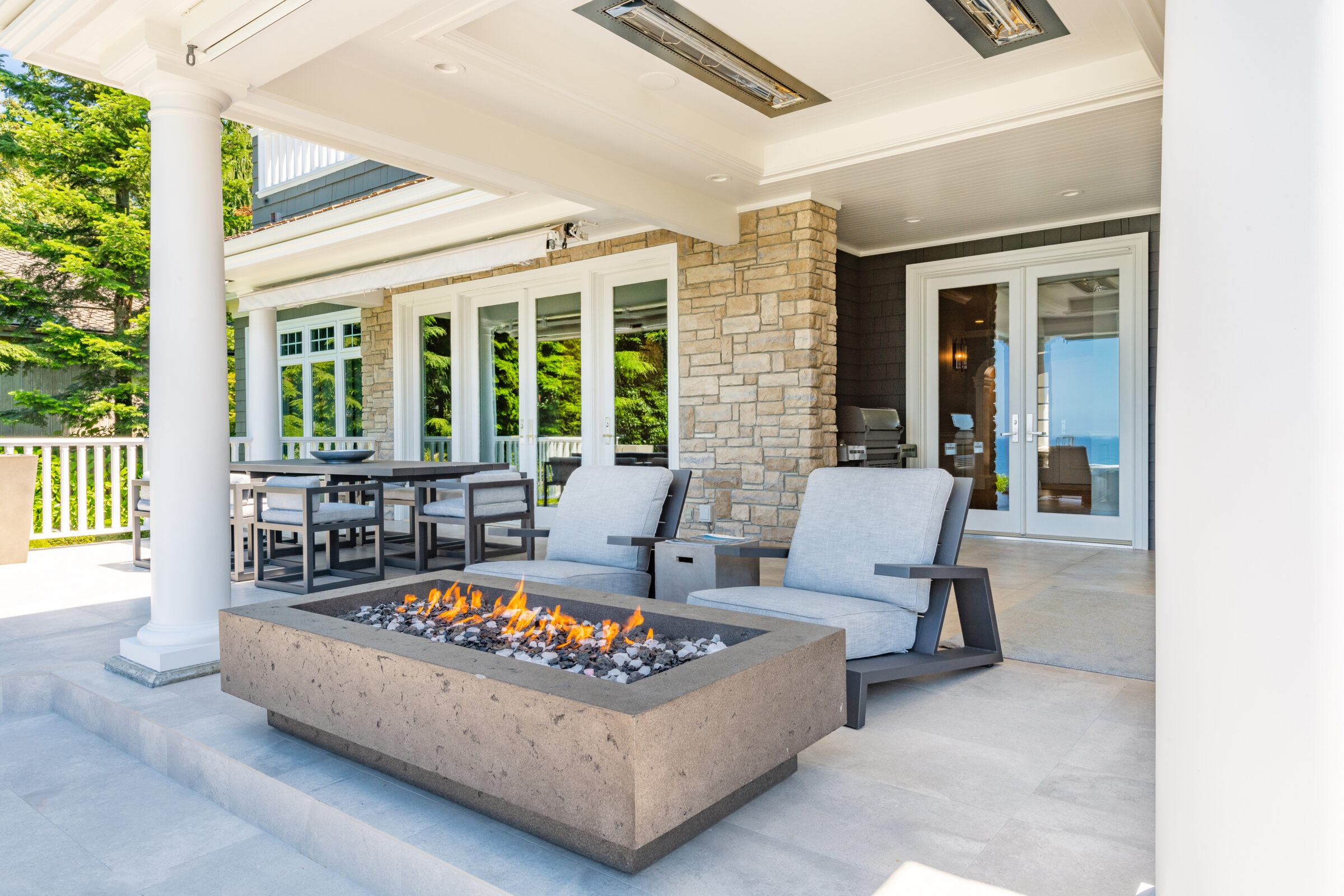 Modern patio with fire pit, comfortable seating, and dining area. Stone facade, large glass doors, lush greenery, and ocean view in the background.