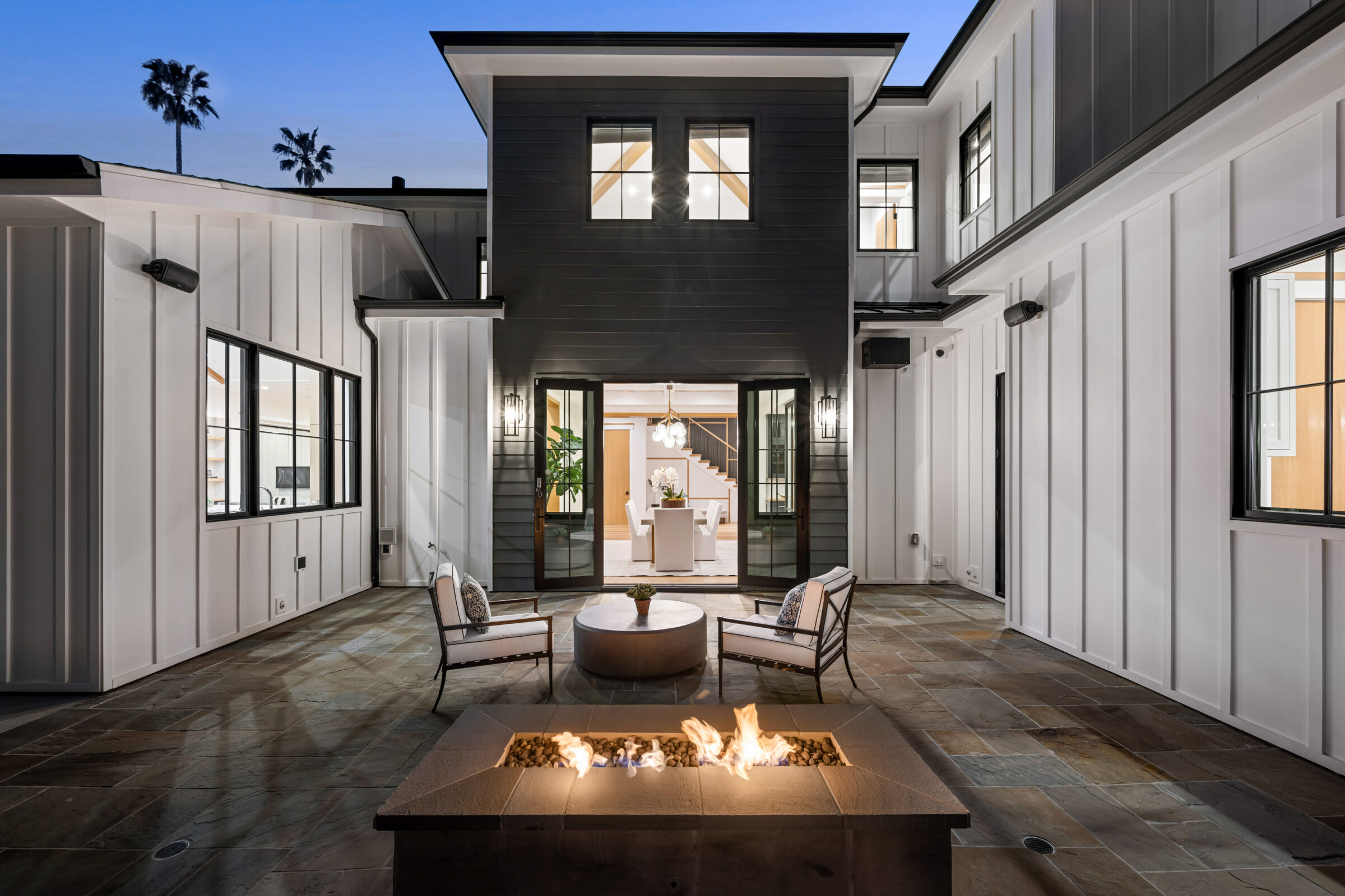 Modern courtyard with fire pit, two chairs, and round table, leading to illuminated dining room. Sleek architecture features large windows and palm trees visible.