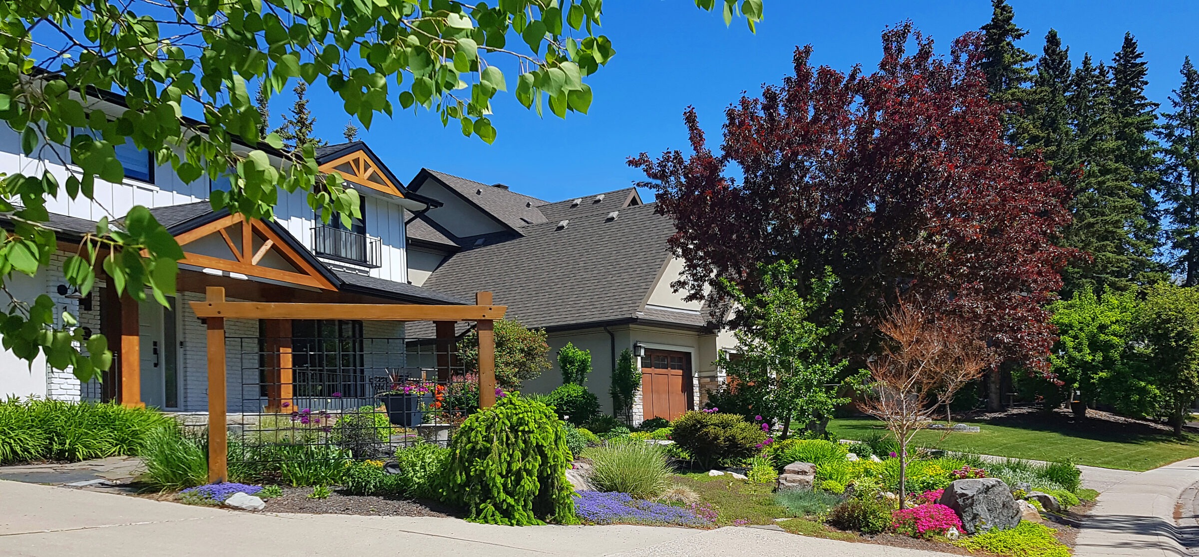 Suburban house with manicured garden, vibrant flowers, and lush trees under a clear blue sky, creating a serene residential scene.