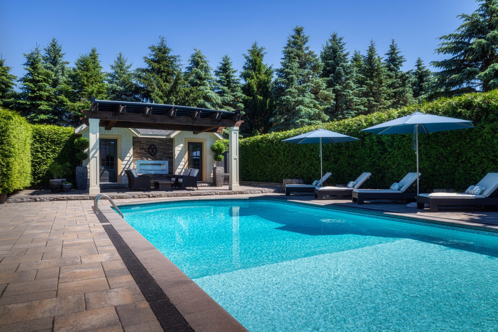 A tranquil backyard features a crystal-clear swimming pool, lounge chairs with umbrellas, and a cozy pavilion, surrounded by lush greenery.