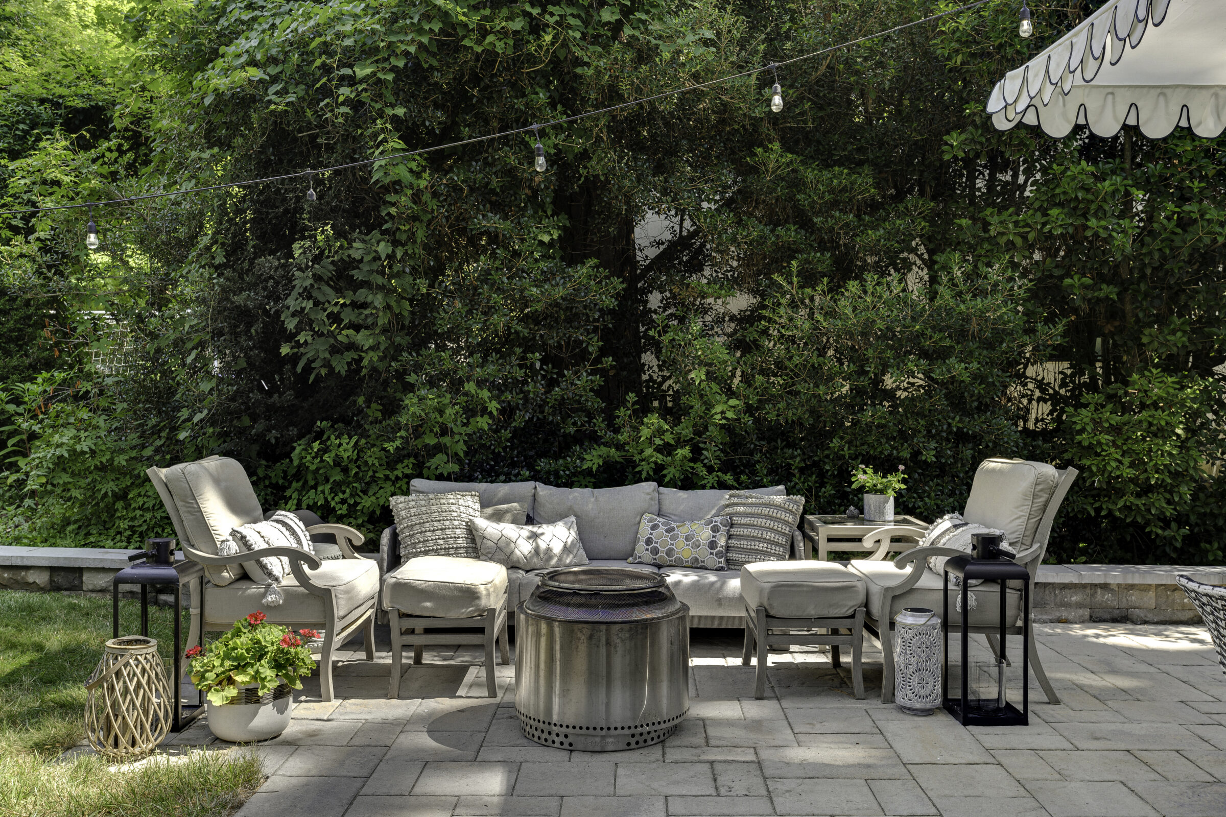 Patio scene with cushioned seating, ottomans, a central fire pit, string lights, surrounded by lush greenery and decorative lanterns on a paved area.