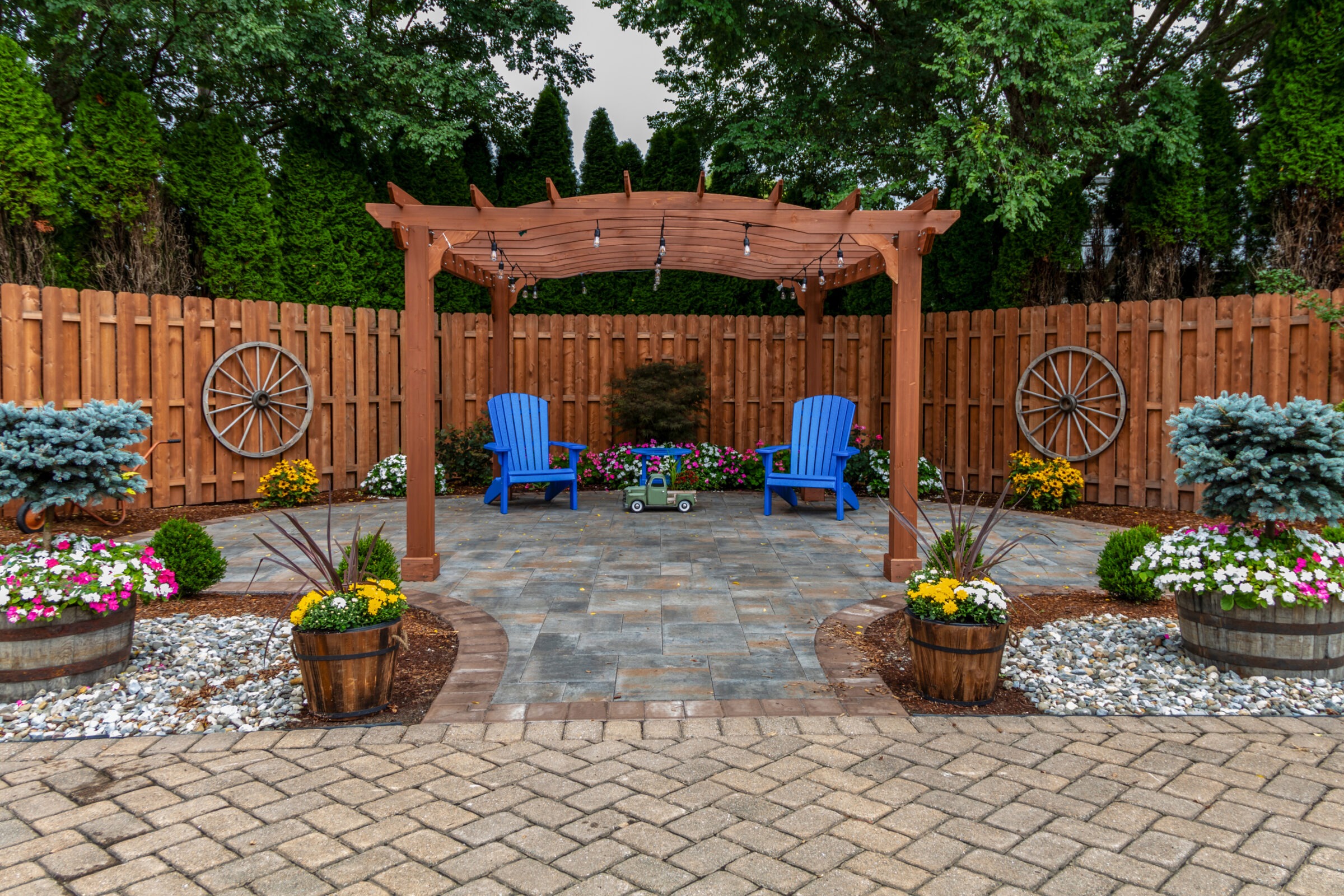 A cozy backyard features a wooden pergola, two blue chairs, decorative wagon wheels, and vibrant flower arrangements, enclosed by a tall wooden fence.