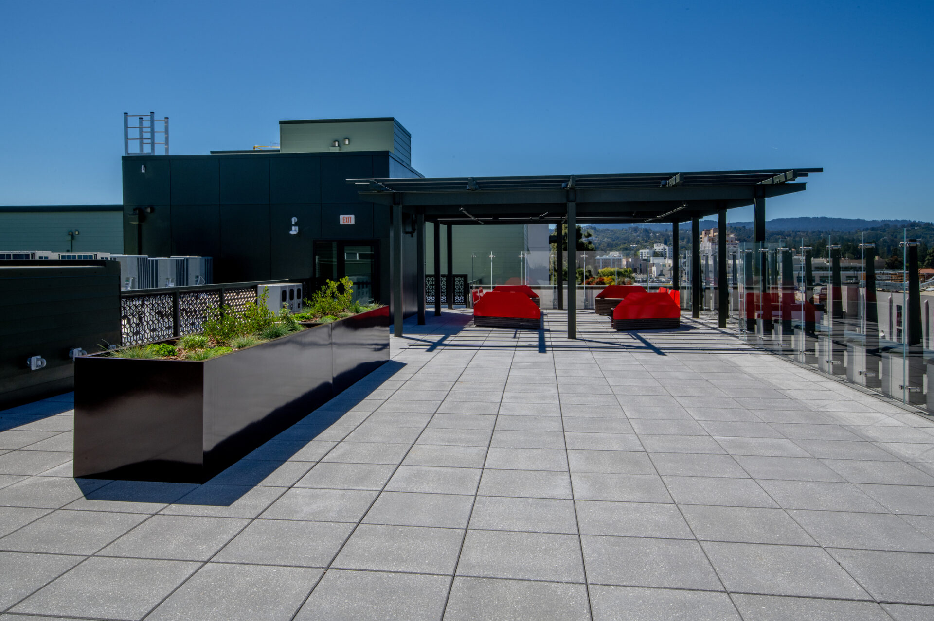 Rooftop terrace with red seating, glass railing, and views of distant hills under clear blue sky. Modern design with planters and shaded area.