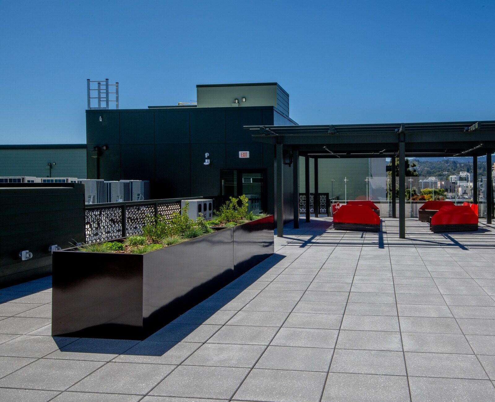 Rooftop terrace with red seating, glass railing, and views of distant hills under clear blue sky. Modern design with planters and shaded area.