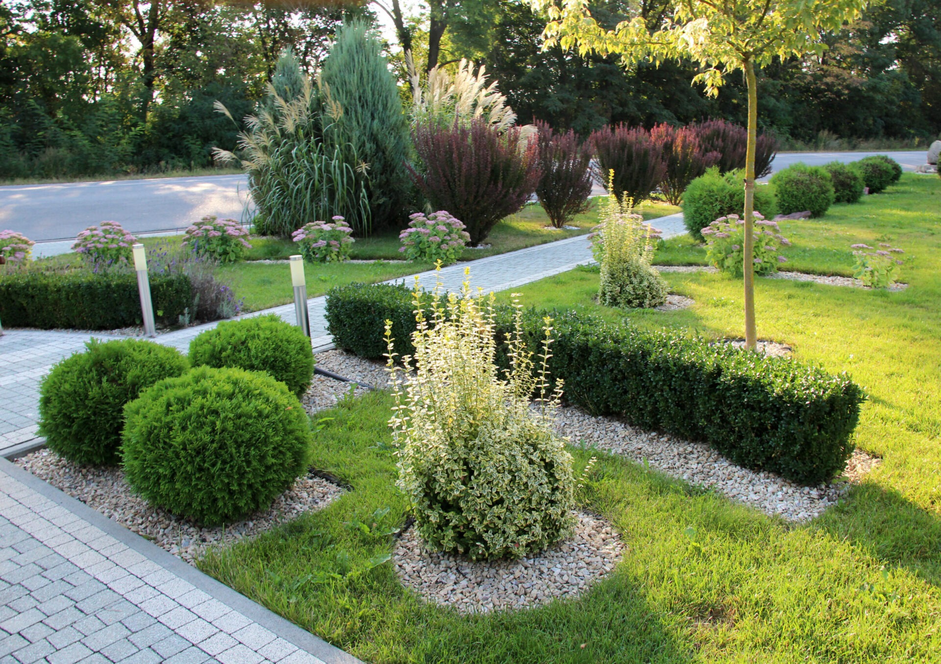 A landscaped garden with neatly trimmed shrubs, trees, and gravel beds, situated along a paved path under a canopy of taller trees.