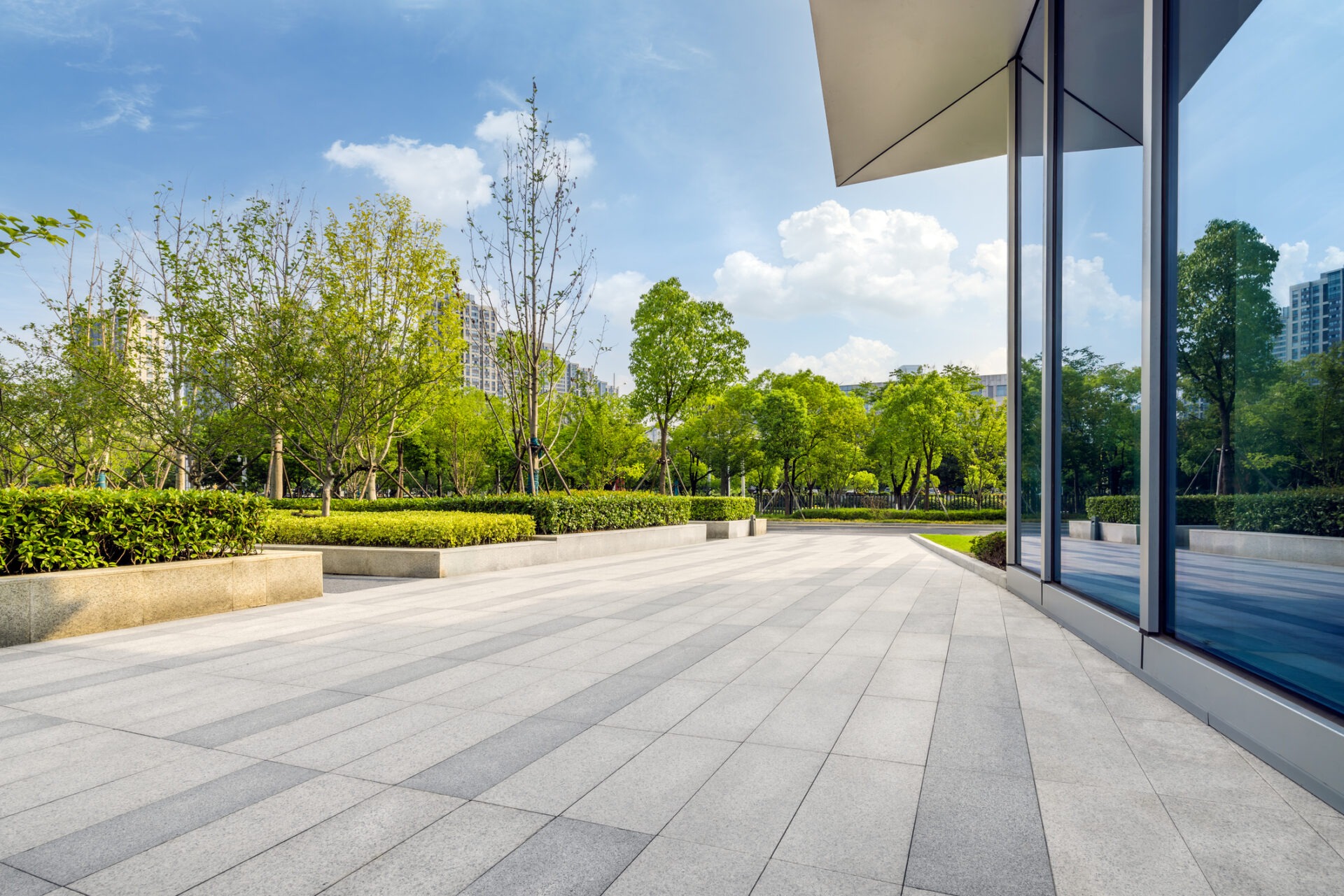 Modern urban park with a sleek building reflecting green trees. Open plaza with patterned tiles extending to landscaped gardens under a bright sky.