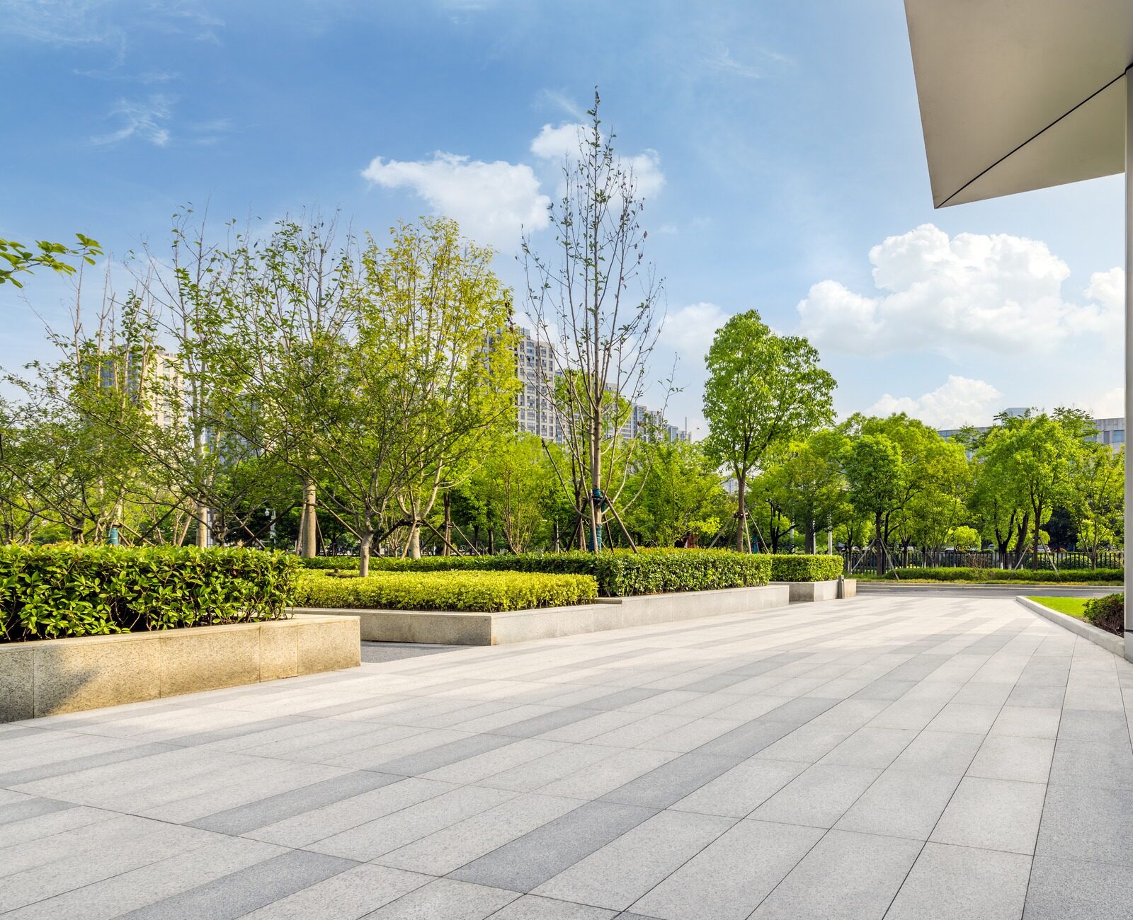 Modern urban park with a sleek building reflecting green trees. Open plaza with patterned tiles extending to landscaped gardens under a bright sky.