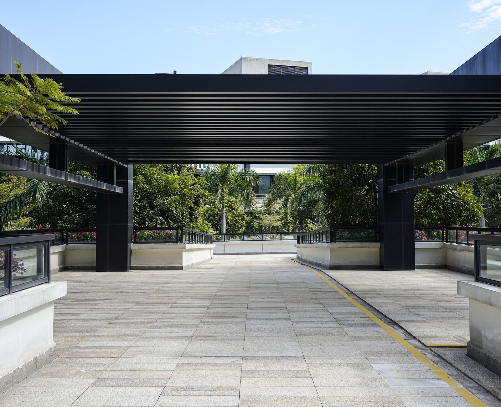Modern architectural walkway with black beams, surrounded by lush greenery and blue sky. No people are present in this serene urban setting.