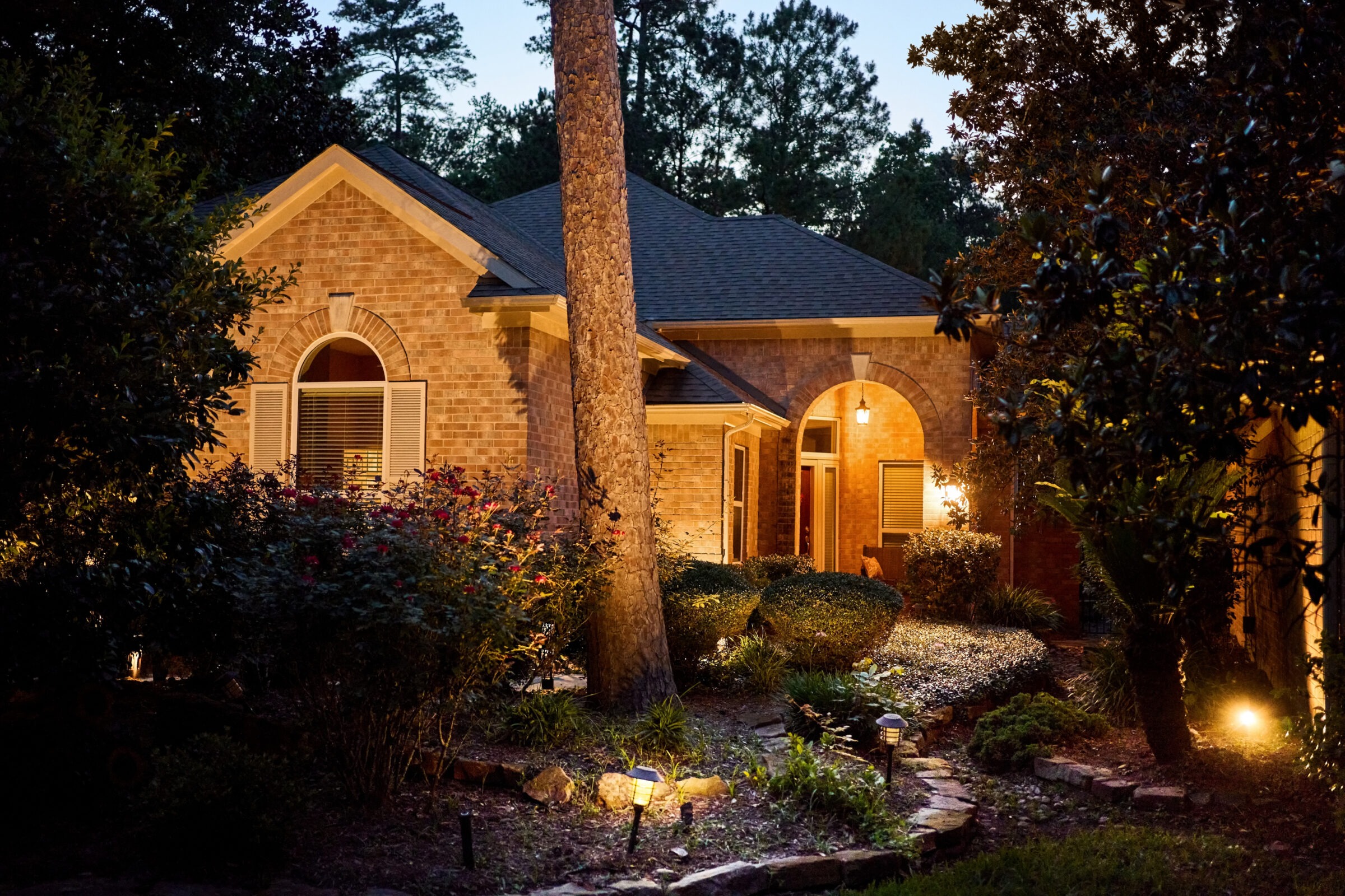 A cozy brick house illuminated at dusk, surrounded by manicured bushes and trees, creating a peaceful, inviting atmosphere in a suburban setting.
