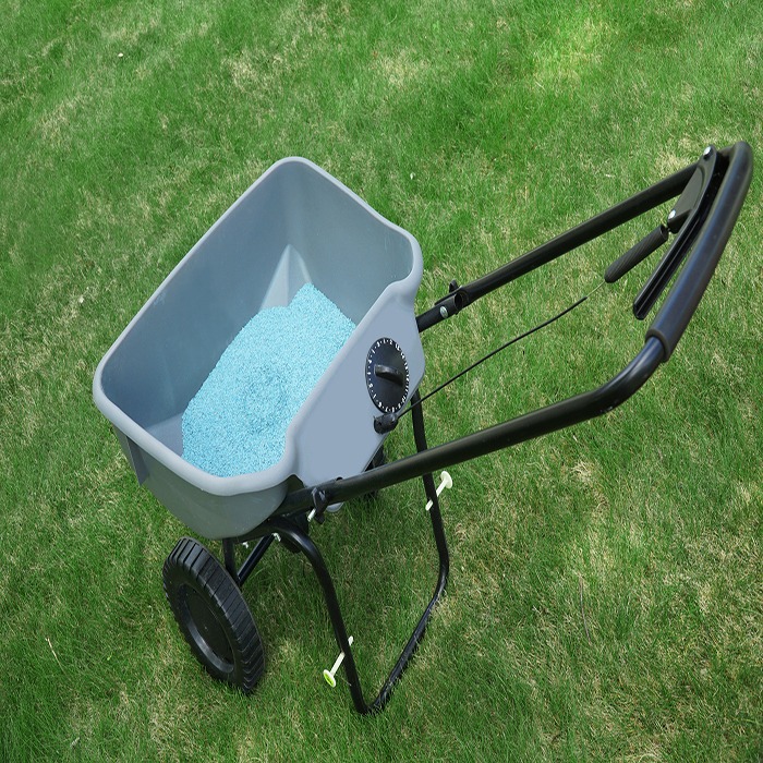 A lawn spreader on green grass is filled with blue granules, designed for evenly distributing materials like seeds or fertilizer in a garden.