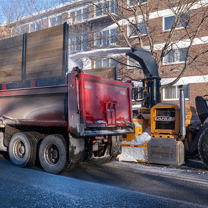 A red dump truck and snow removal machine operating near a brick building on a snowy urban street under clear blue skies.