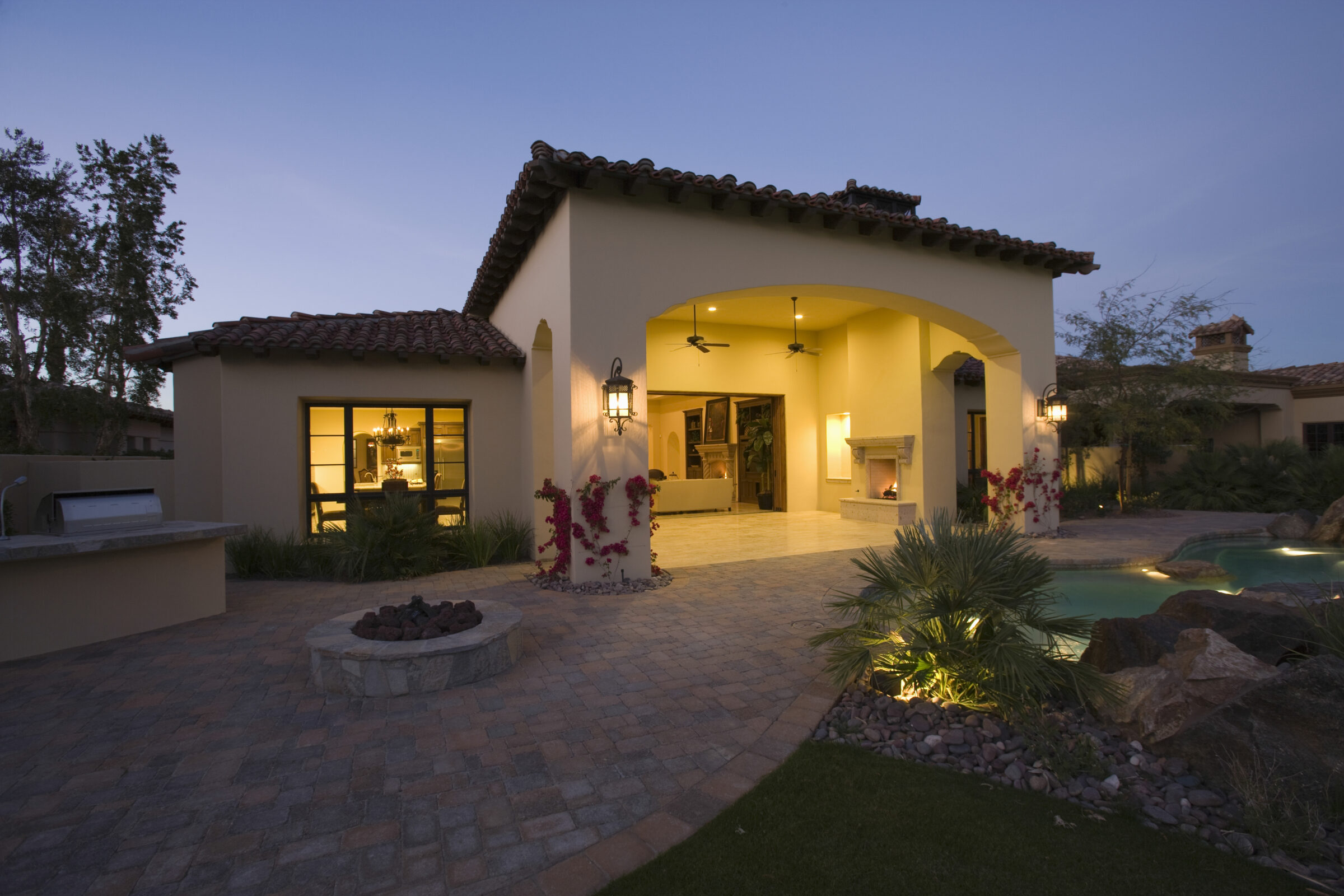 This image depicts a Mediterranean-style house at dusk, featuring a pool, patio area, and ornate lighting, surrounded by desert landscaping.