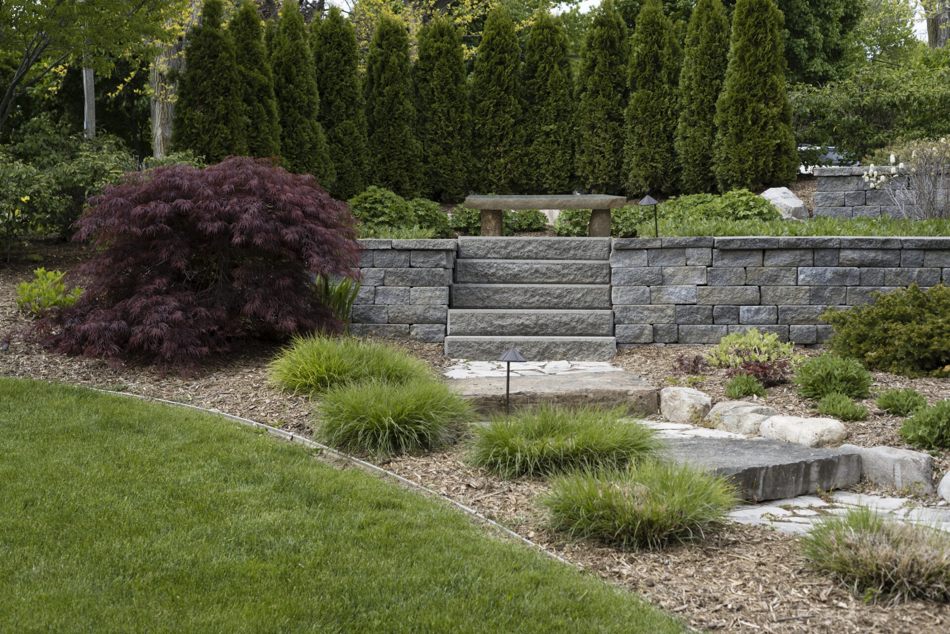 Stone steps lead through a landscaped garden with lush greenery, bushes, and a stone bench framed by neatly arranged trees.