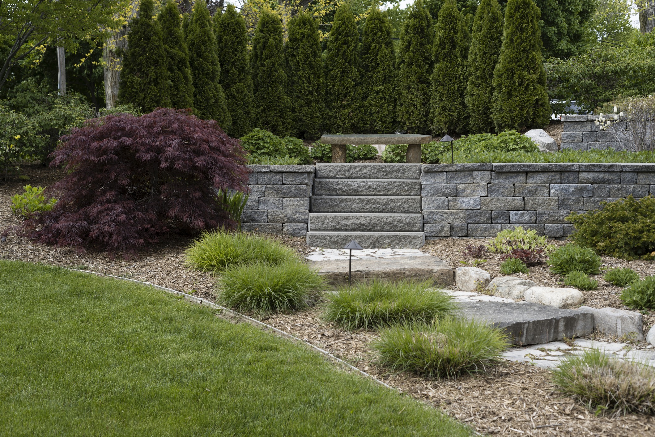 Stone steps lead through a landscaped garden with lush greenery, bushes, and a stone bench framed by neatly arranged trees.
