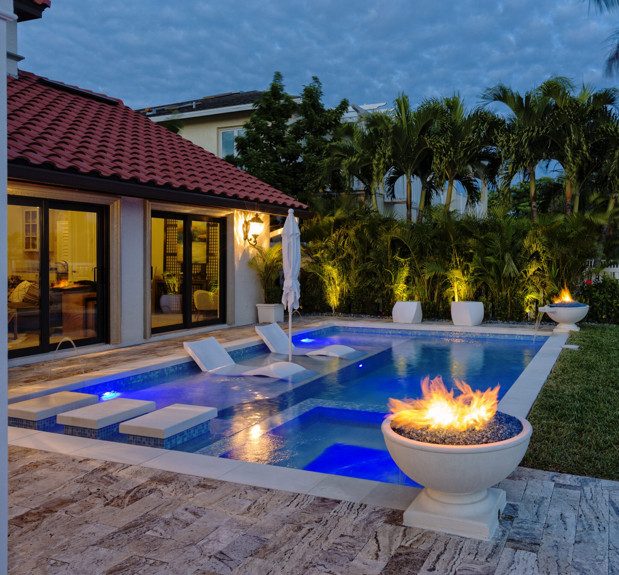 A modern backyard features a serene pool with loungers, fire bowls, and lush greenery under a twilight sky. Warm lighting illuminates the scene.