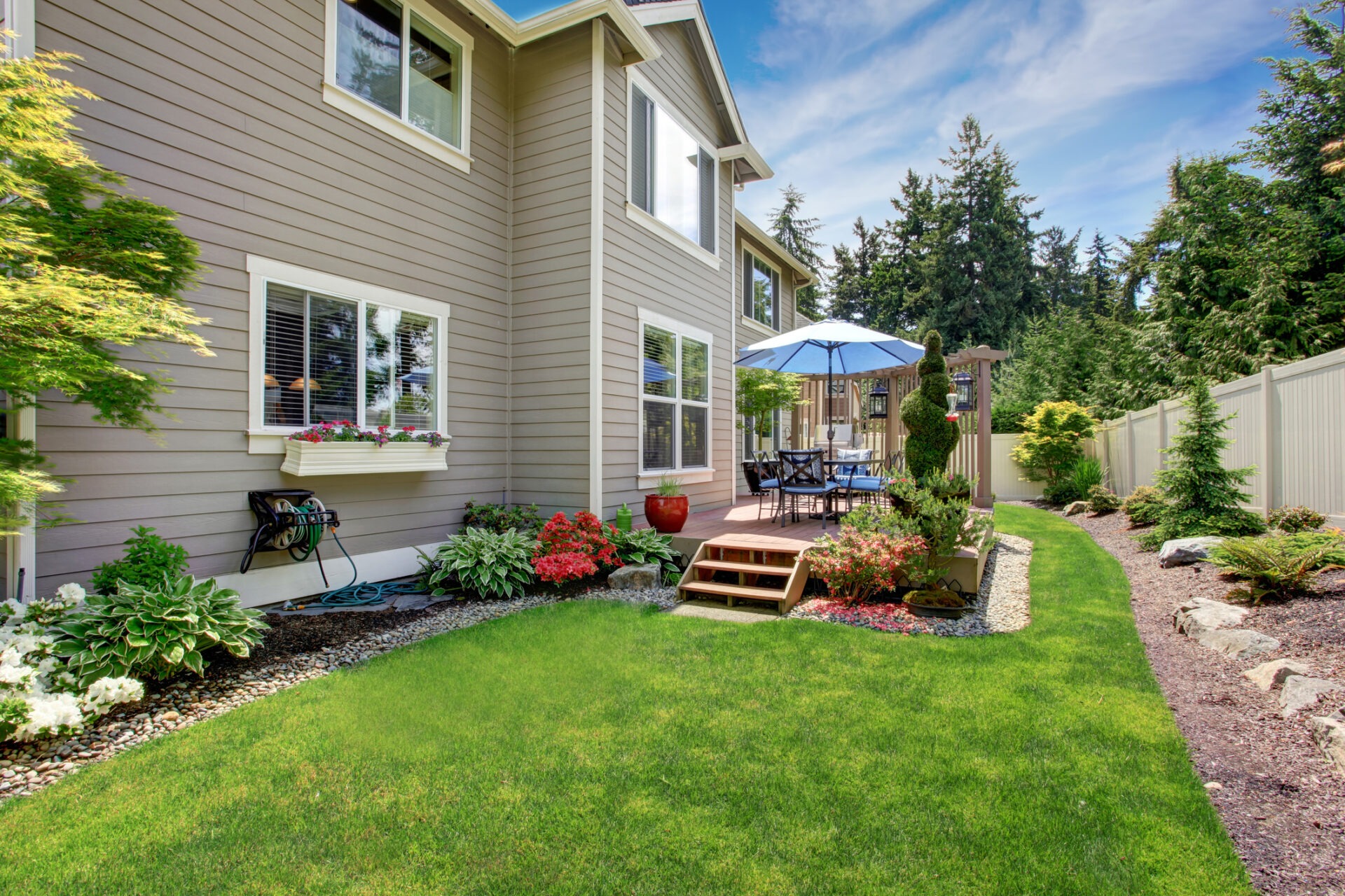 House with beige siding, a landscaped backyard, deck with blue umbrella, outdoor dining set, and surrounding trees under a clear sky.