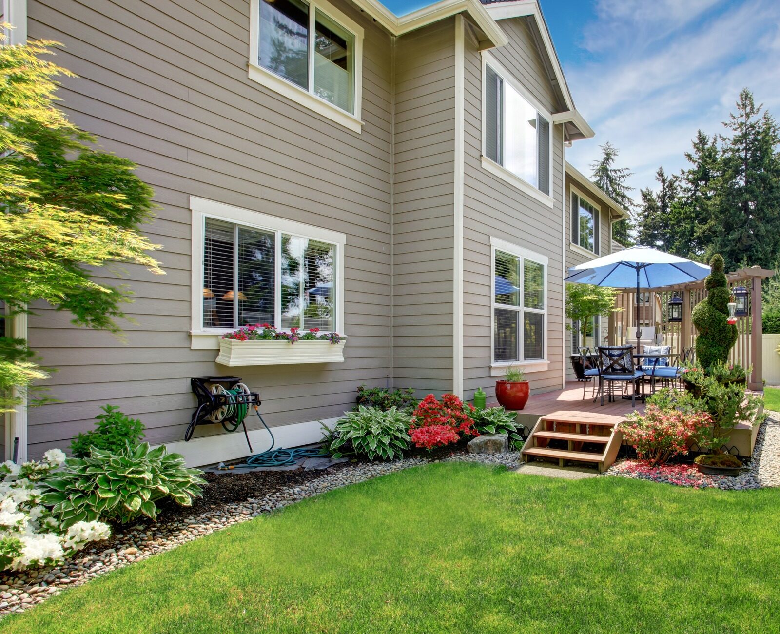 House with beige siding, a landscaped backyard, deck with blue umbrella, outdoor dining set, and surrounding trees under a clear sky.