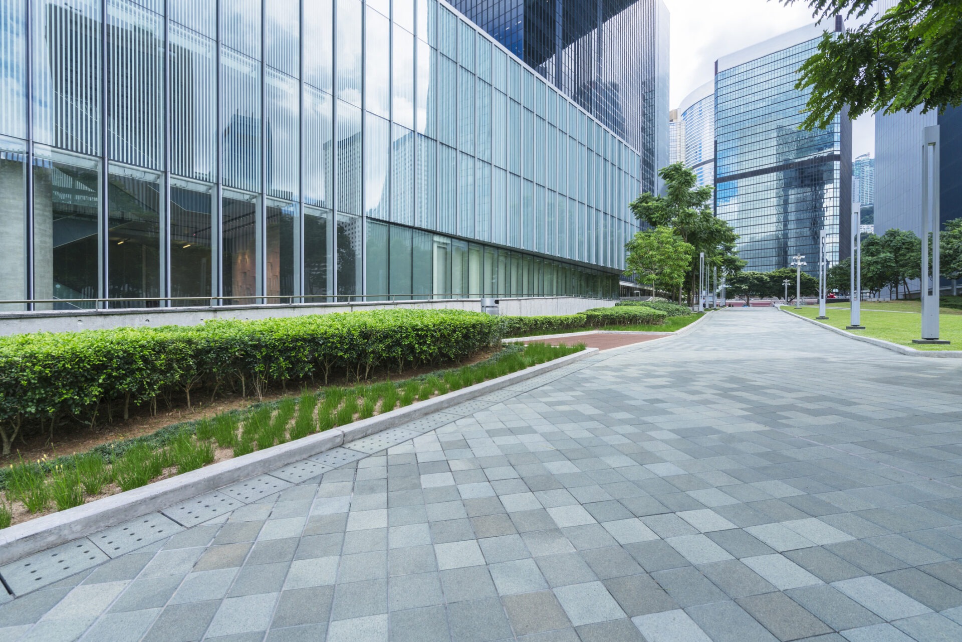 Modern urban scene with glass skyscrapers, clean tiled walkway, greenery, and overcast sky. Quiet area, suggesting a professional business district environment.
