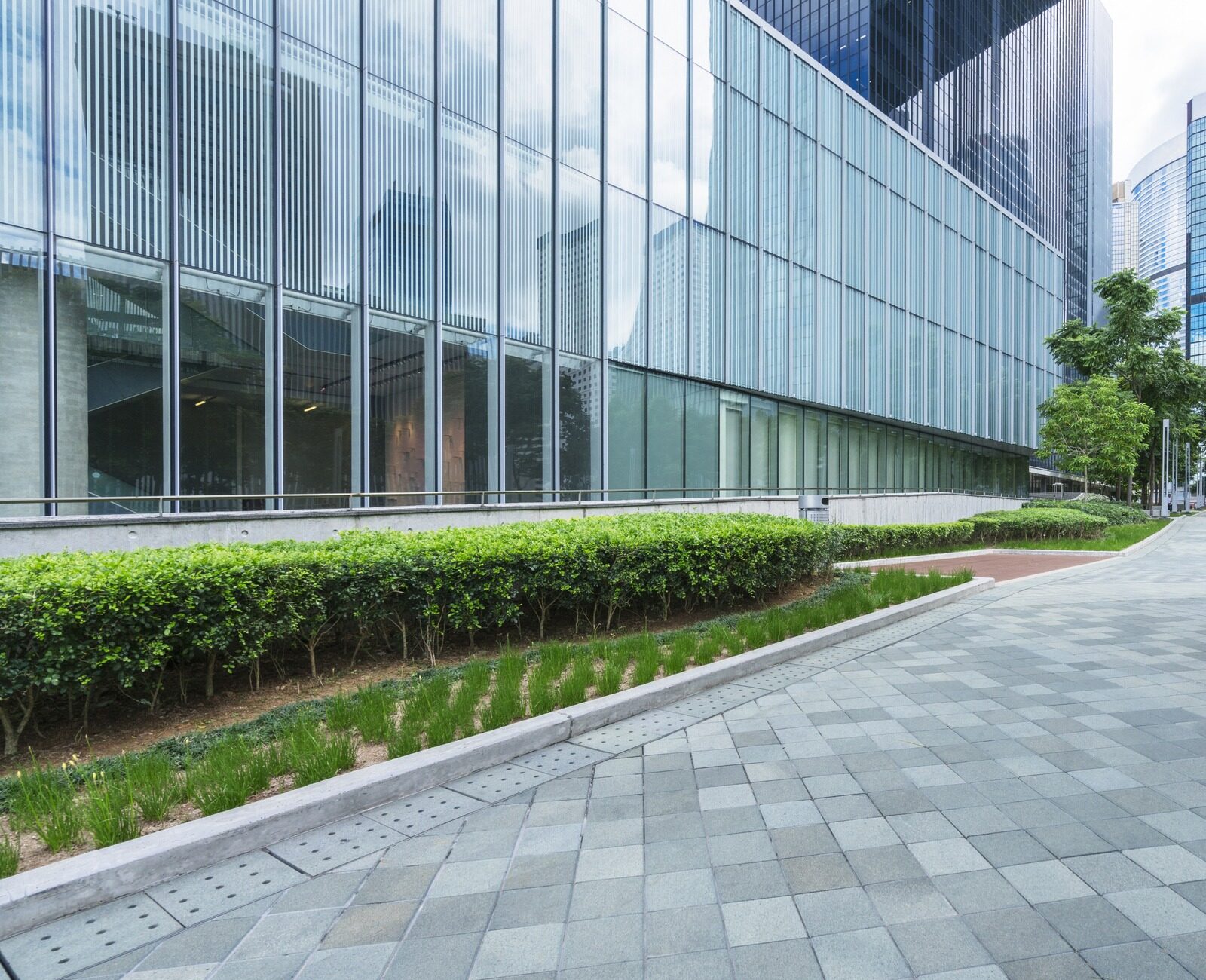 Modern urban scene with glass skyscrapers, clean tiled walkway, greenery, and overcast sky. Quiet area, suggesting a professional business district environment.