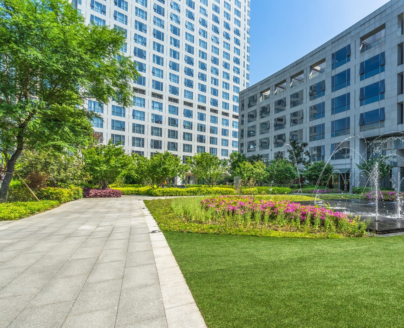 Modern courtyard with tall buildings, lush green garden, and a central fountain. Pathway leads through vibrant plants under a clear blue sky.