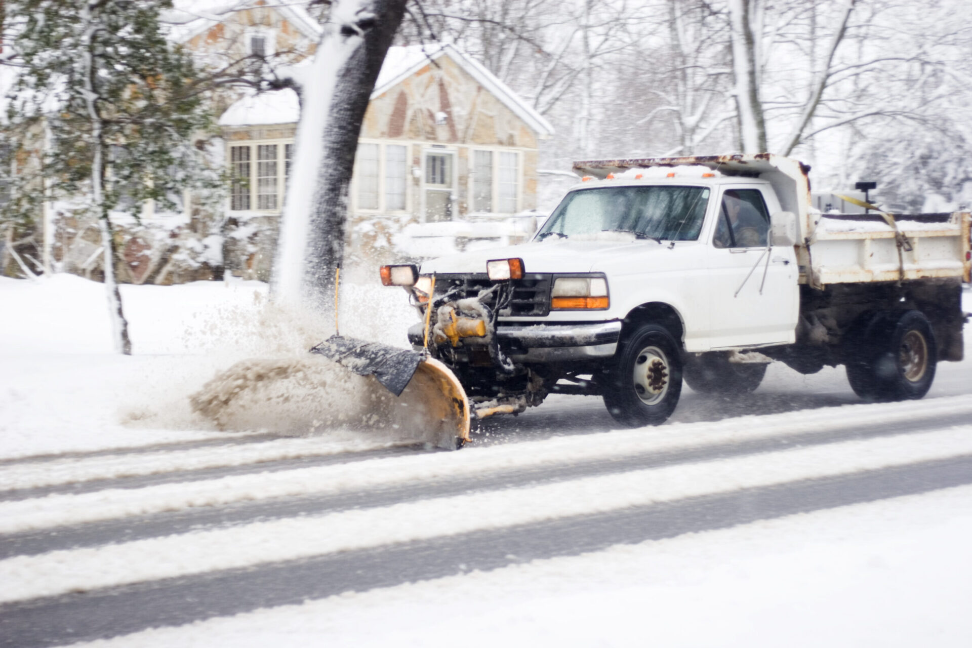 A snowplow truck clears a snowy road in a residential area, passing by a snow-covered house and trees during winter.