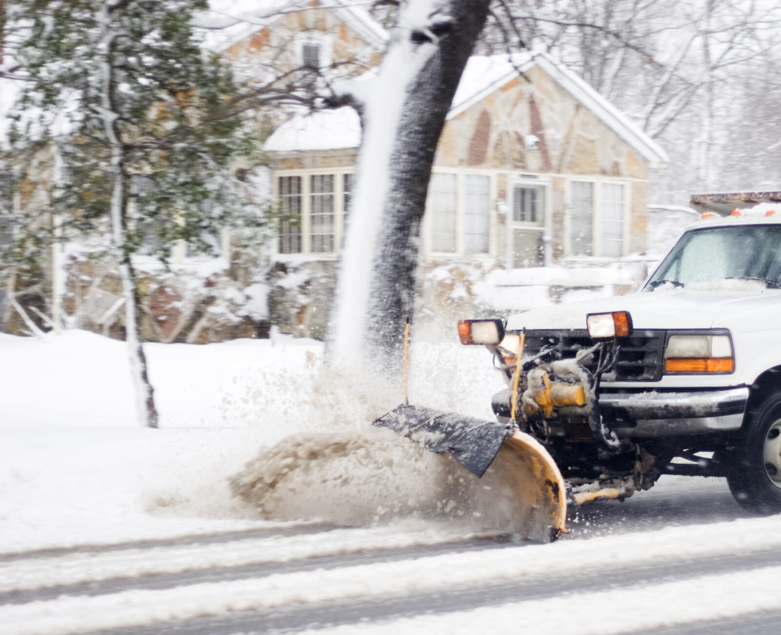 A snowplow truck clears a snowy road in a residential area, passing by a snow-covered house and trees during winter.