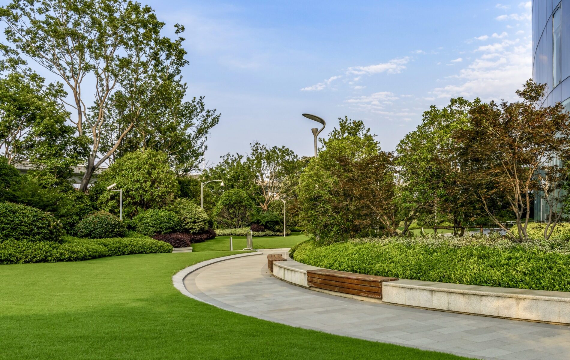 A modern park setting with lush green lawns, curved walkway, trees, and a sleek glass building, under a clear blue sky.