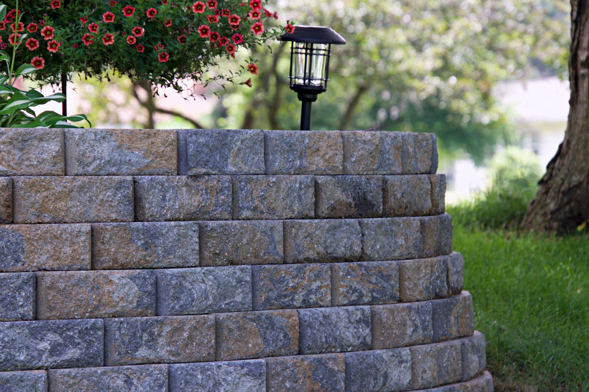 Stone retaining wall with a lantern and vibrant red flowers, surrounded by greenery, under overcast sky, creating a peaceful garden scene.