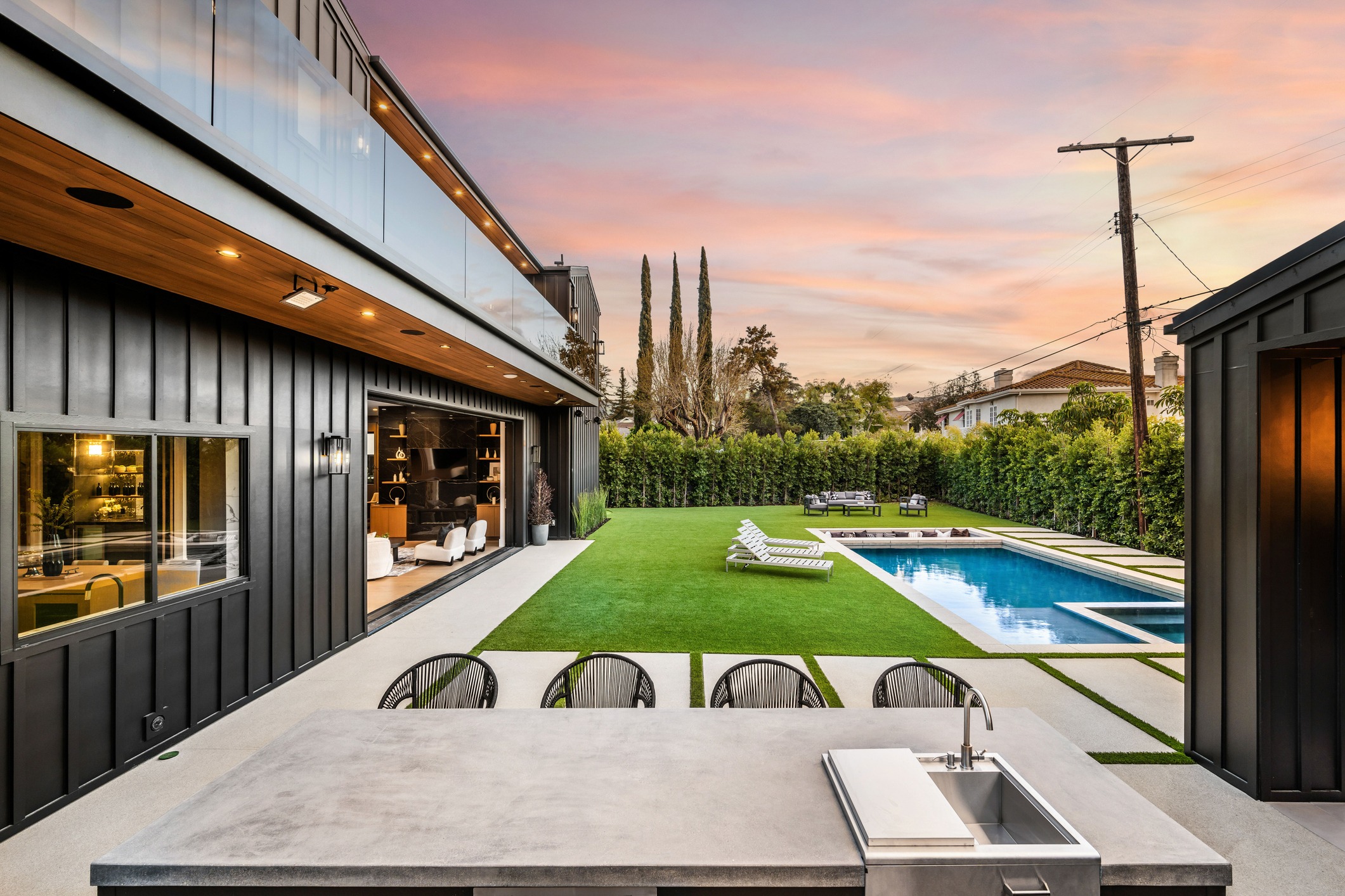 Modern backyard with a swimming pool, outdoor kitchen, and lounge area at sunset.