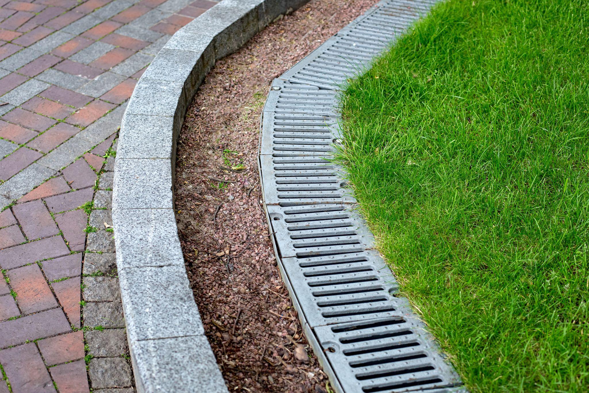 Curved drainage channel alongside brick paver pathway and green lawn